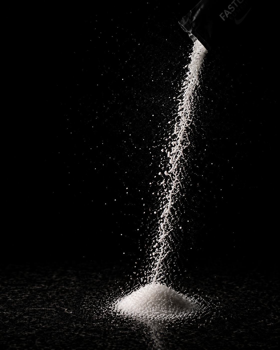 White granulated sugar pouring in a fine stream from a dark container against a solid black background, with a small pile of sugar forming at the bottom.